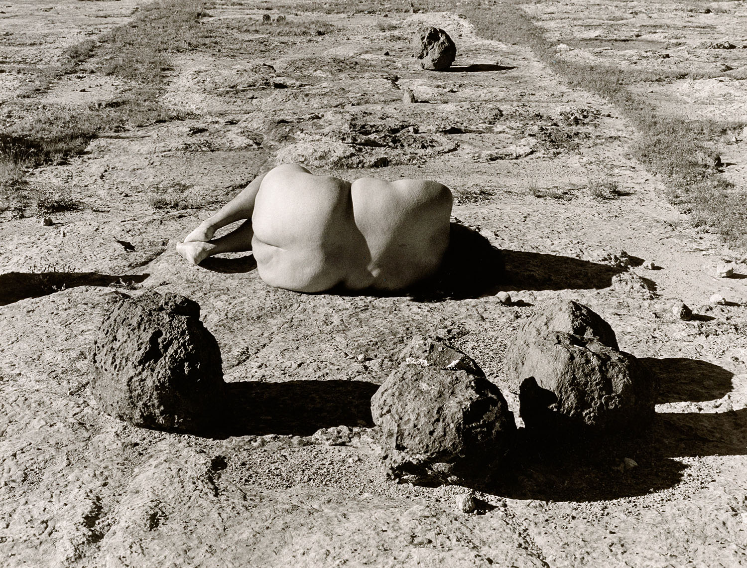 Laura Aguilar print, self portrait of self, lying on dirt, naked on a side, in child pose seen from back with three rocks positioned in the foreground