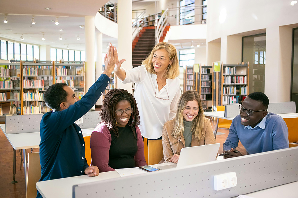 Group of students sitting at desk in front of laptop and advisor looking over smiling and giving high-fiveto one