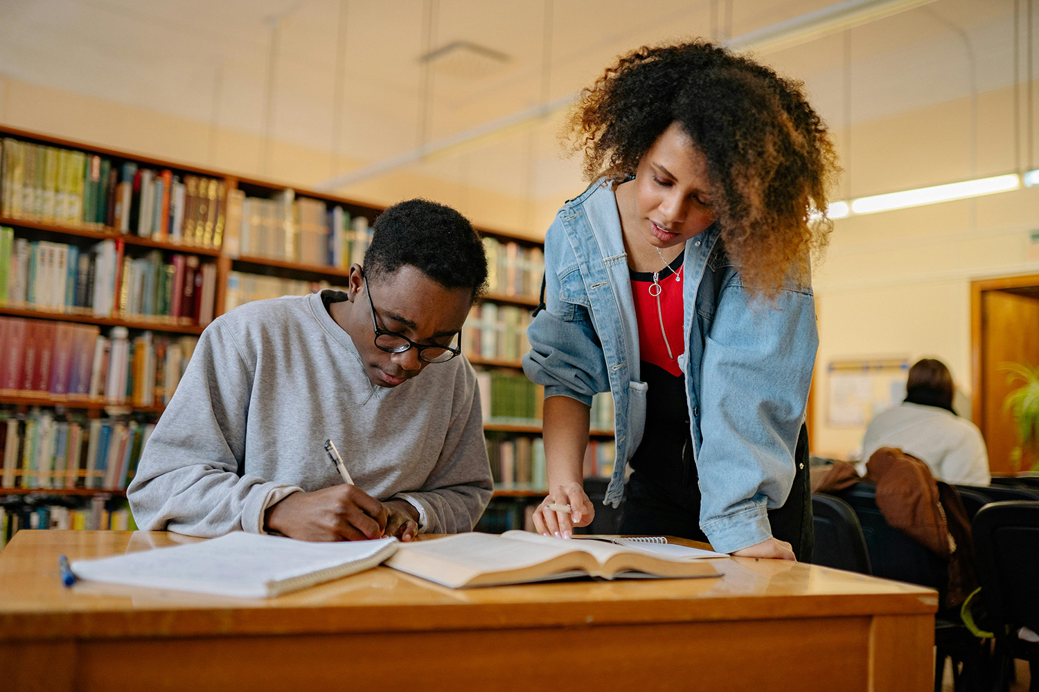 Standing TA leans over student siting at desk in front of books and explains what to do