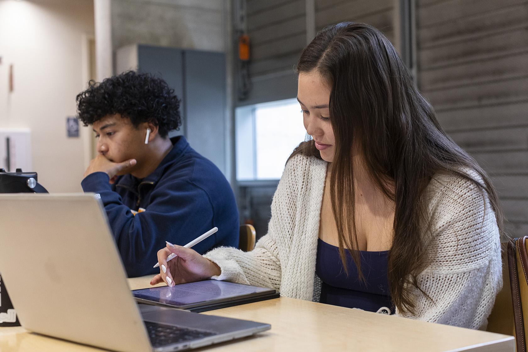 Boy and girl student in UCSB library at desk writing in front of their own laptop