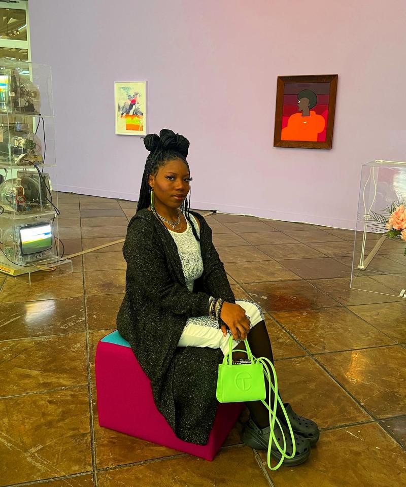 Mariah Webber posing in an art room, sitting on a chair and holding a lime green purse